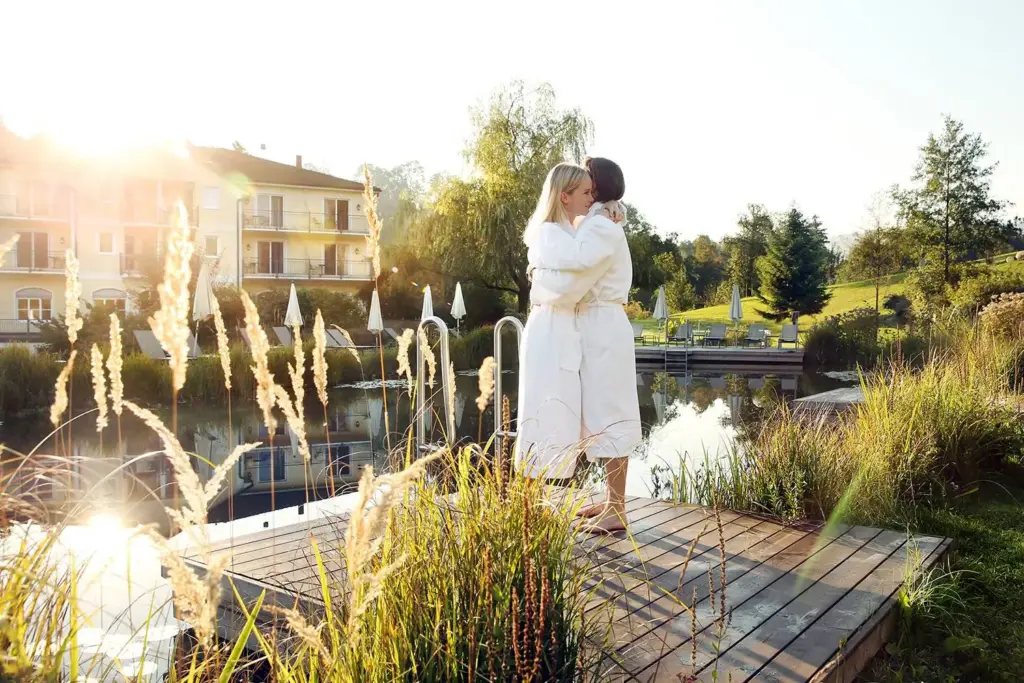 Pärchen umarmt sich in Bademänteln vor einem Schwimmteich.